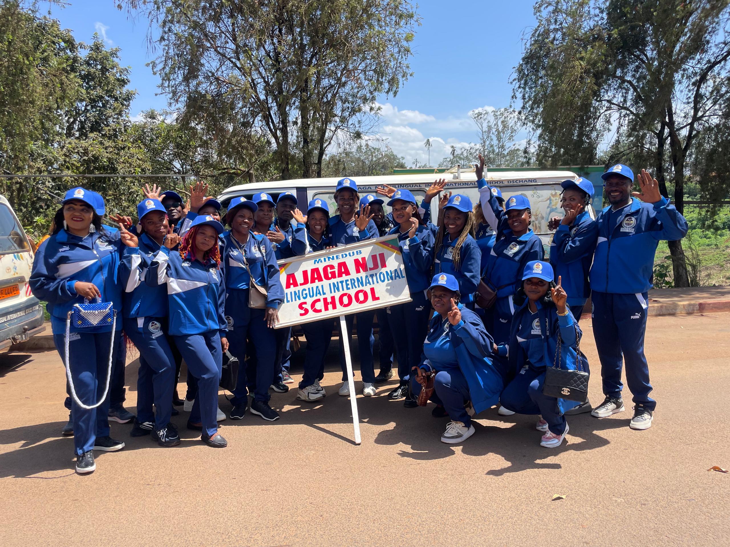 Ajaga Nji school group in blue tracksuits waving beside the school sign
